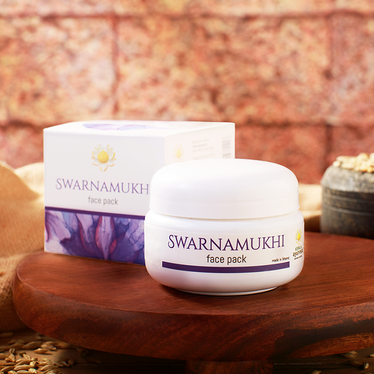 A white jar of Kerala Ayurveda Swarnamukhi Face Pack and its box, positioned on a wooden platter against a brick wall background, with some dry leaves visible. – Kerala Ayurveda India